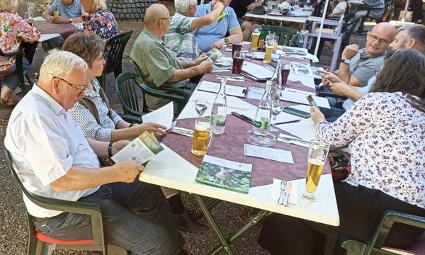 Mehrere Personen sitzen an einem Tisch im Freien, lesen Broschüren und unterhalten sich. Auf dem Tisch stehen Getränke, darunter Bier und Wasserflaschen. Im Hintergrund sind weitere Tische sichtbar.