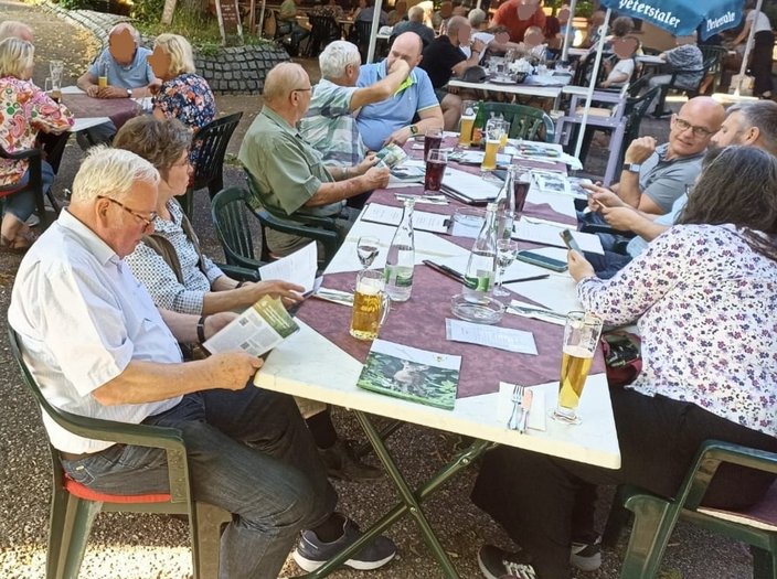 Mehrere Personen sitzen an einem Tisch im Freien, lesen Broschüren und unterhalten sich. Auf dem Tisch stehen Getränke, darunter Bier und Wasserflaschen. Im Hintergrund sind weitere Tische sichtbar.