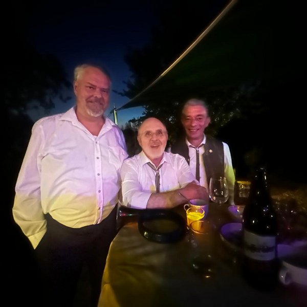 Drei Männer stehen lachend an einem Tisch im Freien bei Nacht. Auf dem Tisch stehen Gläser, eine Flasche und Kerzen. Die Umgebung ist dunkel, möglicherweise ein Garten oder Park.