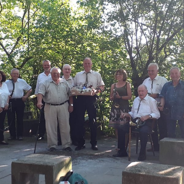 Eine Gruppe von Personen steht, einige mit Musikinstrumenten in den Händen, draußen auf einer gepflasterten Fläche, umgeben von grünen Bäumen und Sonnenschein.