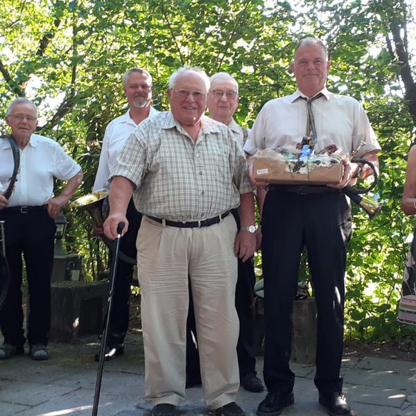 Eine Gruppe älterer Menschen posiert mit Hörnern und einem Geschenkkorb in einem bewaldeten Park. Sie lächeln fröhlich und stehen auf einem gepflasterten Weg unter grünen Bäumen.