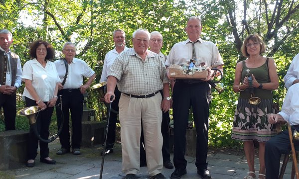 Eine Gruppe älterer Menschen posiert mit Hörnern und einem Geschenkkorb in einem bewaldeten Park. Sie lächeln fröhlich und stehen auf einem gepflasterten Weg unter grünen Bäumen.