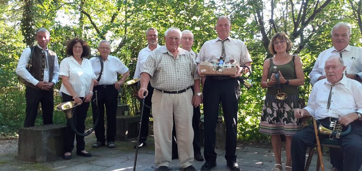 Eine Gruppe älterer Menschen posiert mit Hörnern und einem Geschenkkorb in einem bewaldeten Park. Sie lächeln fröhlich und stehen auf einem gepflasterten Weg unter grünen Bäumen.