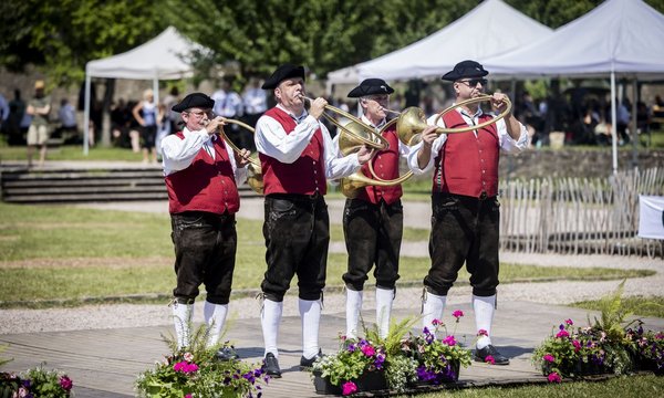 Vier Männer spielen Blasinstrumente; sie sind in roten Westen und dunklen Hüten gekleidet, stehen auf einer hölzernen Bühne im Freien, umgeben von Blumen und Pavillons.
