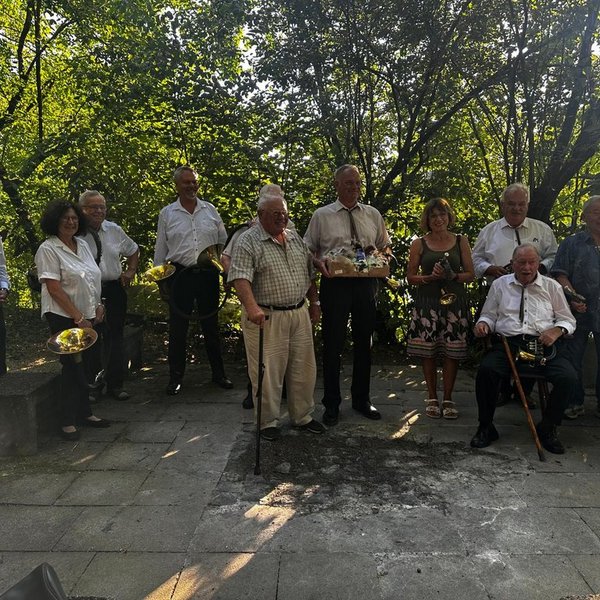 Eine Gruppe älterer Menschen steht oder sitzt im Freien auf einer gepflasterten Fläche, teilweise mit Musikinstrumenten in den Händen, umgeben von dichtem, grünem Laubwerk.