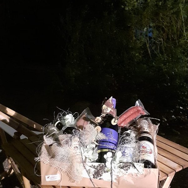 Ein Präsentkorb mit Weinflasche, Wurst und Bier steht auf einem Holztisch. Draußen, im Halbdunkel, umgeben von Bäumen und blauem Abendhimmel.