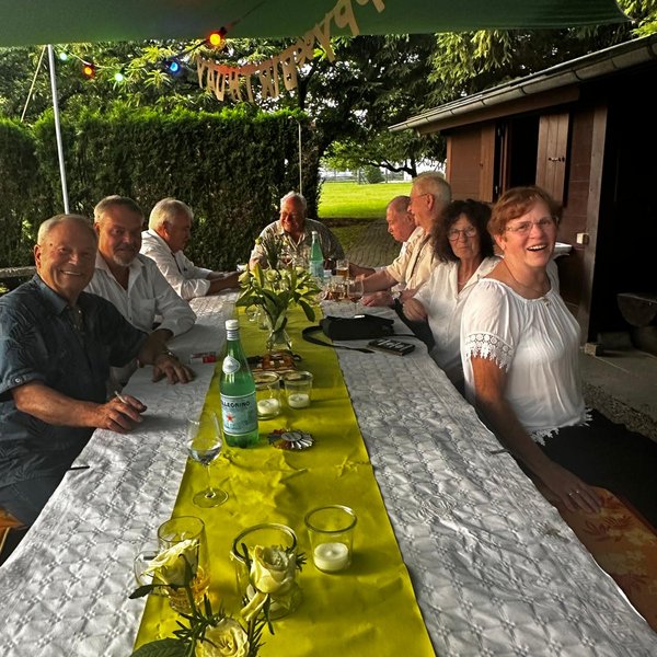 Menschen sitzen an einem festlich gedeckten, langen Tisch unter einem Pavillon. Fröhliche Stimmung mit Getränken und Dekoration. „Happy Birthday“-Girlande hängt. Garten und Hütte im Hintergrund.